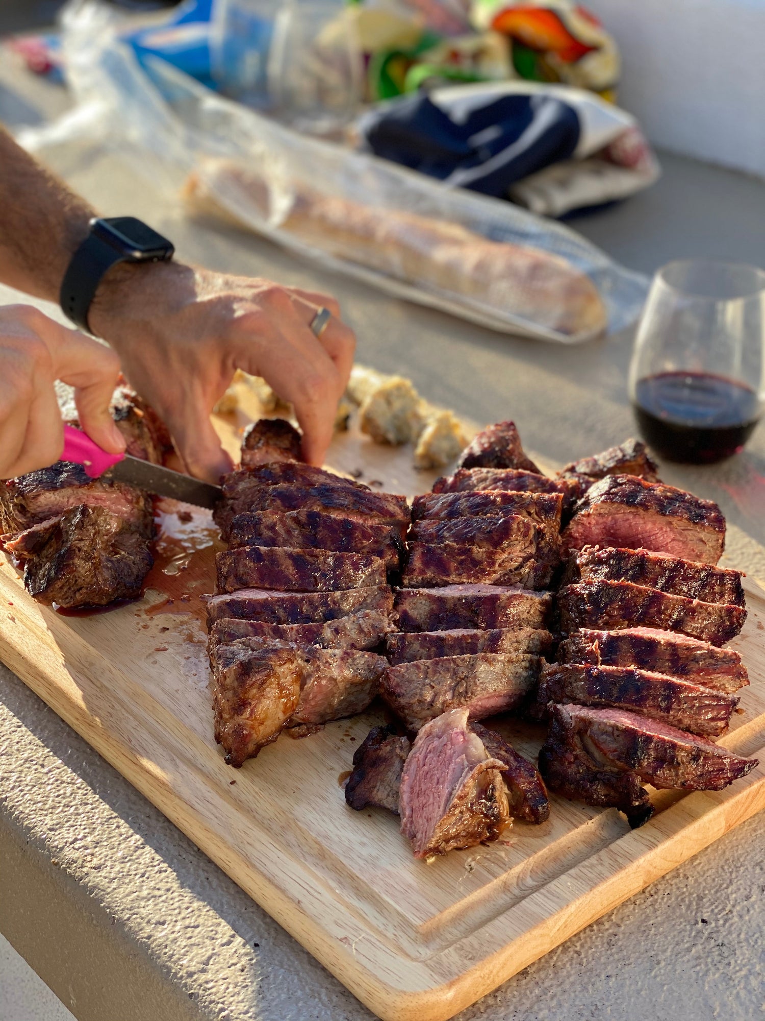 Sliced grilled steaks on a wooden cutting board with hands carving pieces and a glass of red wine nearby