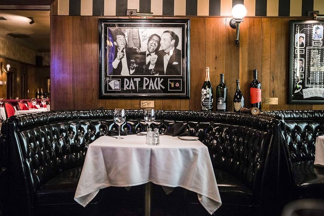 Leather banquette with white-tablecloth table set for two under framed 'RAT PACK' photo and wine bottles on wood-paneled wall