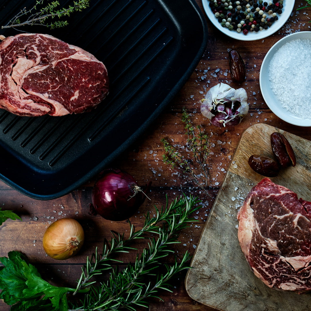 Two raw ribeye steaks beside a cast-iron grill pan, rosemary, onions, garlic, coarse salt and peppercorns on a wooden board