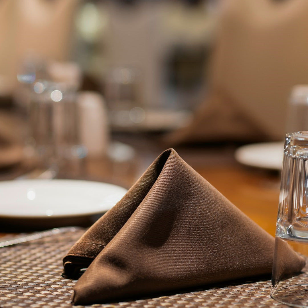 Brown cloth napkin folded into a pyramid on a woven placemat beside an upside-down water glass and a blurred plate.