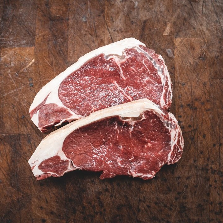 Two raw ribeye steaks with fat cap on a well-scratched wooden cutting board.