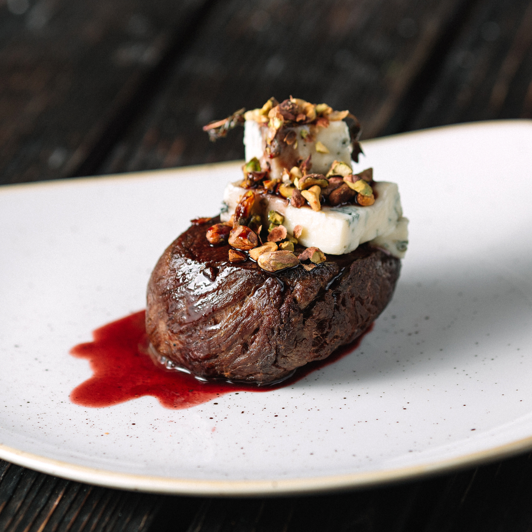 Seared beef filet topped with gorgonzola cubes, chopped pistachios and herbs, with red-wine sauce on a white plate