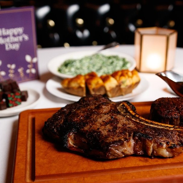 Grilled bone-in steak on a wooden board with sides, a lit candle, and a purple card reading Happy Mother's Day