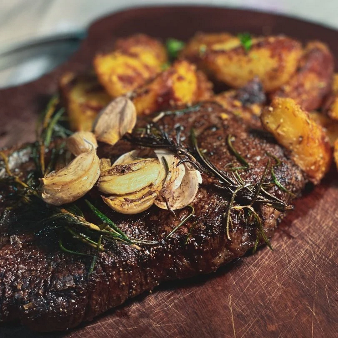 Pan-seared ribeye steak on a cutting board topped with roasted garlic and rosemary, with roasted potatoes behind