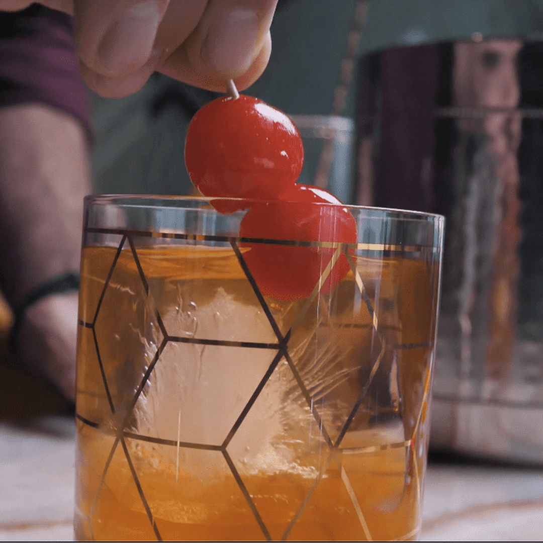 Old Fashioned-style cocktail in patterned glass with large clear ice cube, two cocktail cherries on a pick placed on top.