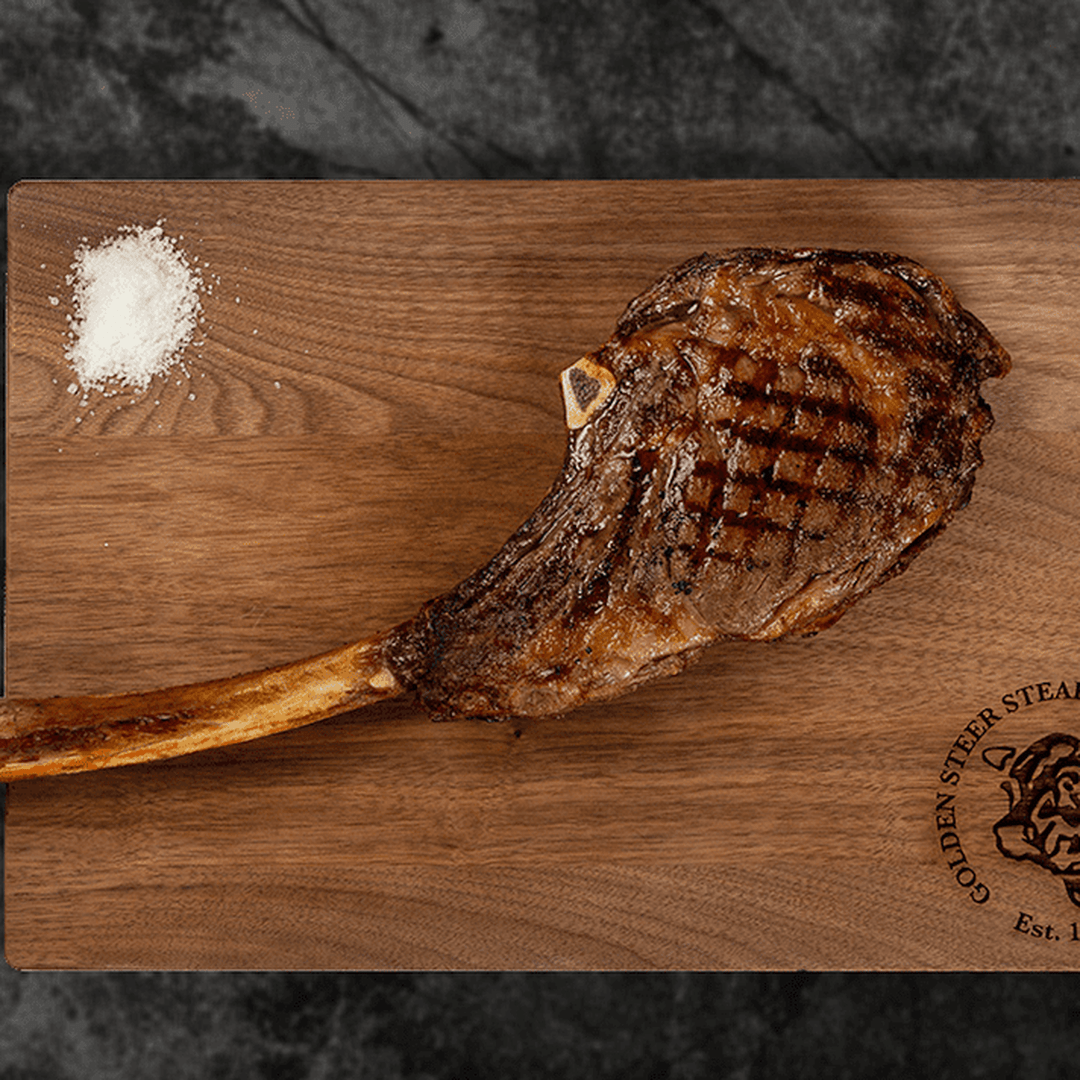 Grilled tomahawk steak with long bone on wooden cutting board, coarse salt pile, stamped 'GOLDEN STEER STEAK' 'Est.'