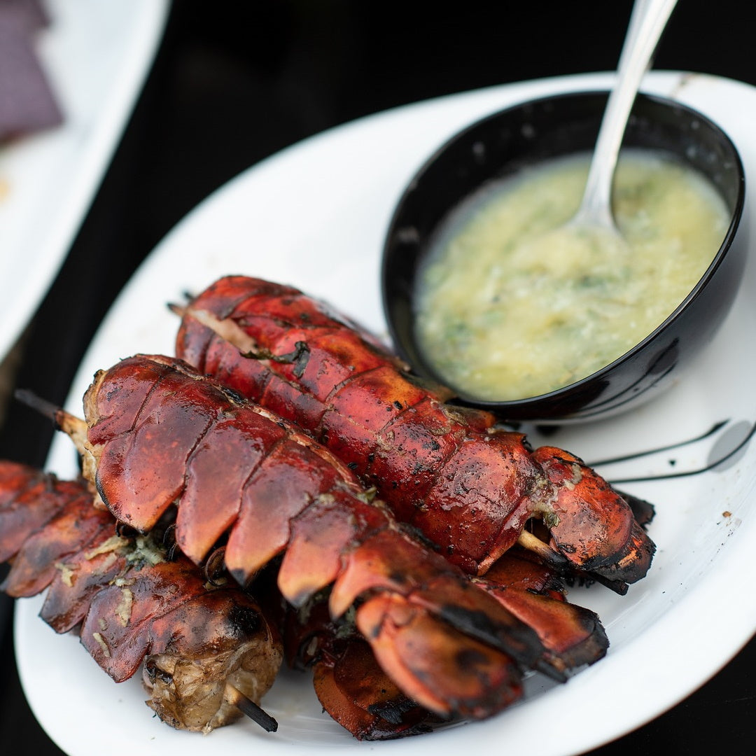 Grilled lobster tails on skewers, charred and glazed, served with a bowl of melted garlic-butter sauce on a white plate.