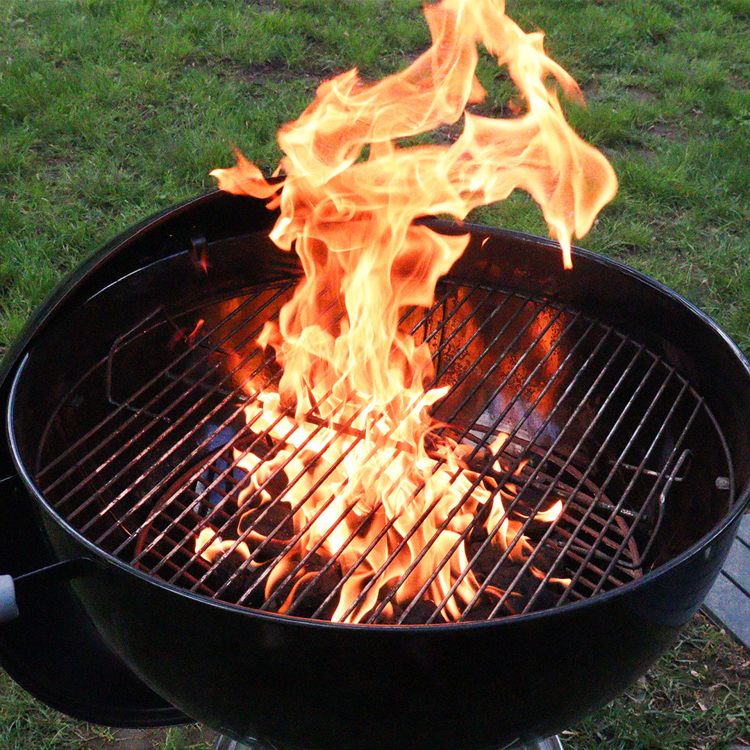 Charcoal kettle grill with tall orange flames over hot coals, set on backyard grass.