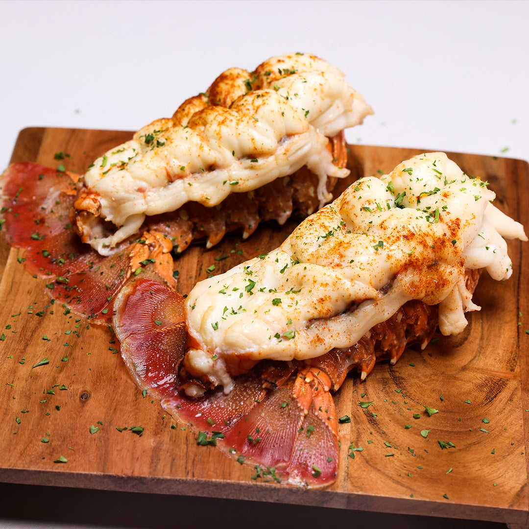 Two broiled lobster tails sprinkled with chopped parsley and paprika on a wooden serving board