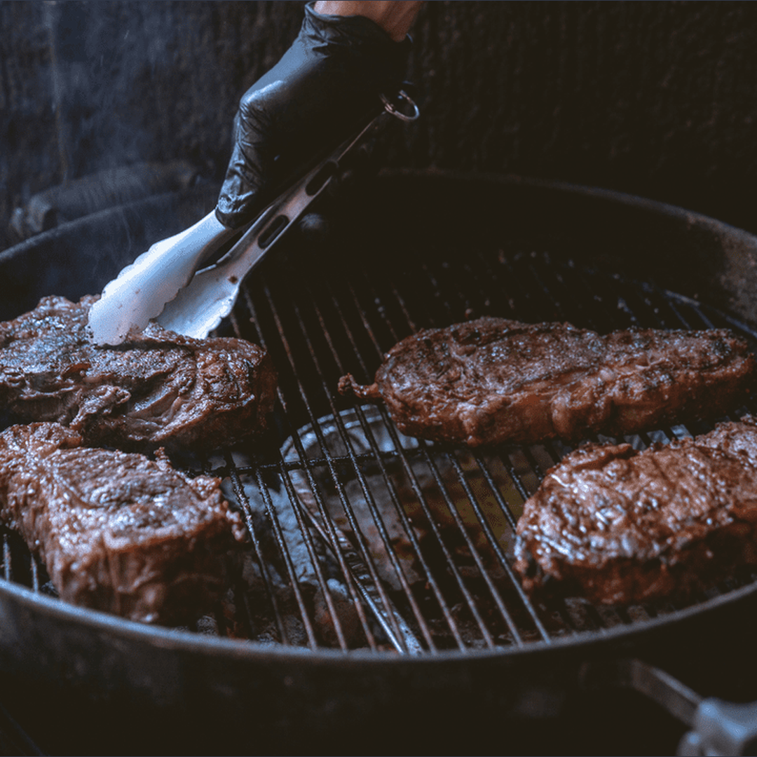 Four steaks searing on a charcoal grill while a gloved hand lifts one with metal tongs