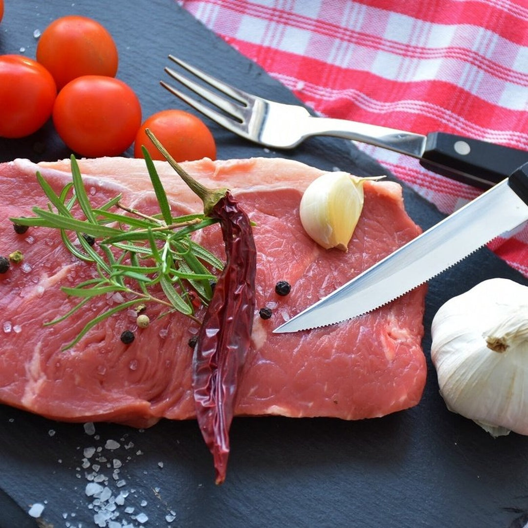 Raw steak on slate with rosemary, dried chili, garlic, peppercorns, coarse salt, cherry tomatoes, fork and serrated knife
