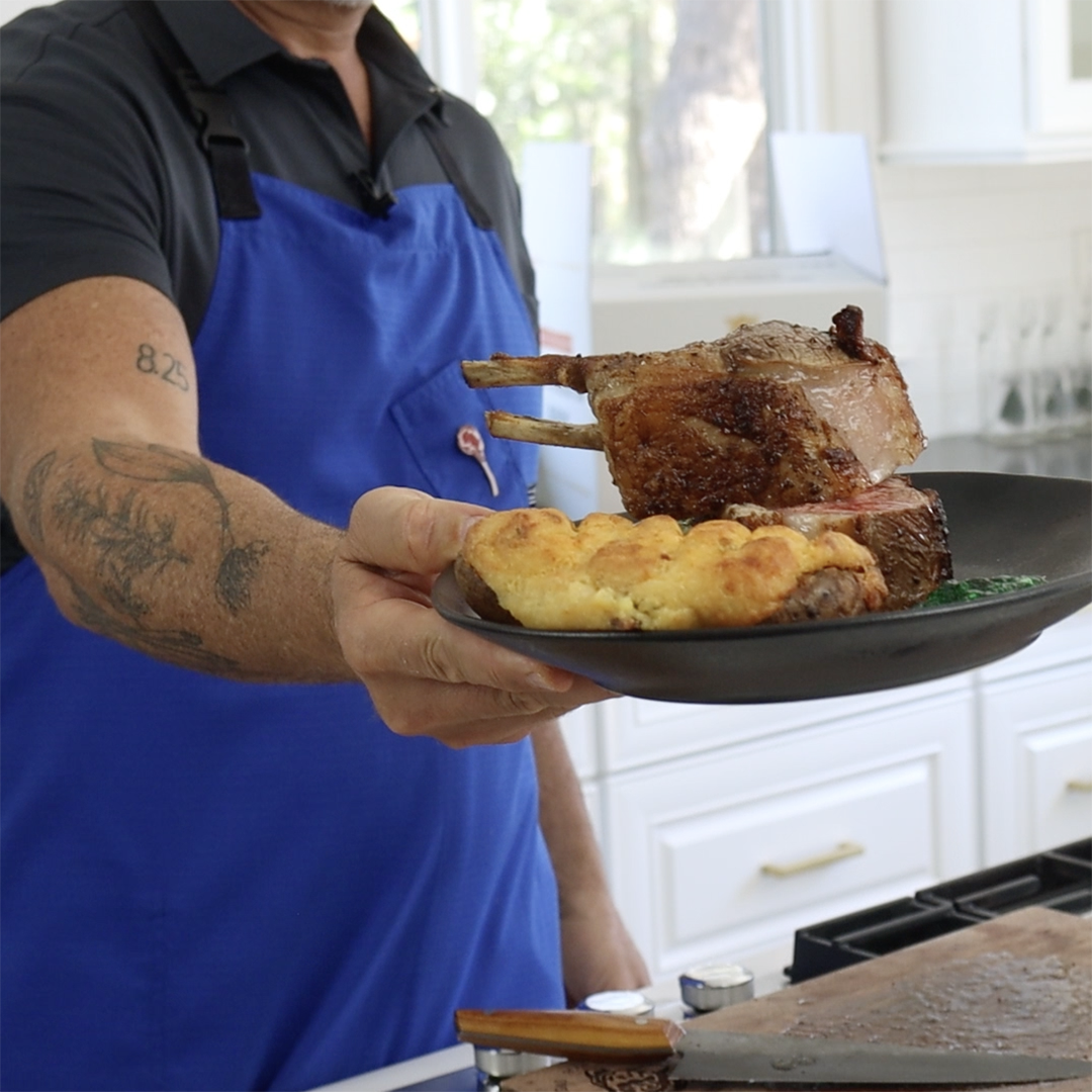 Cook holding a plate with double-cut rack of lamb and twice-baked potato; forearm tattoo reads 8.25