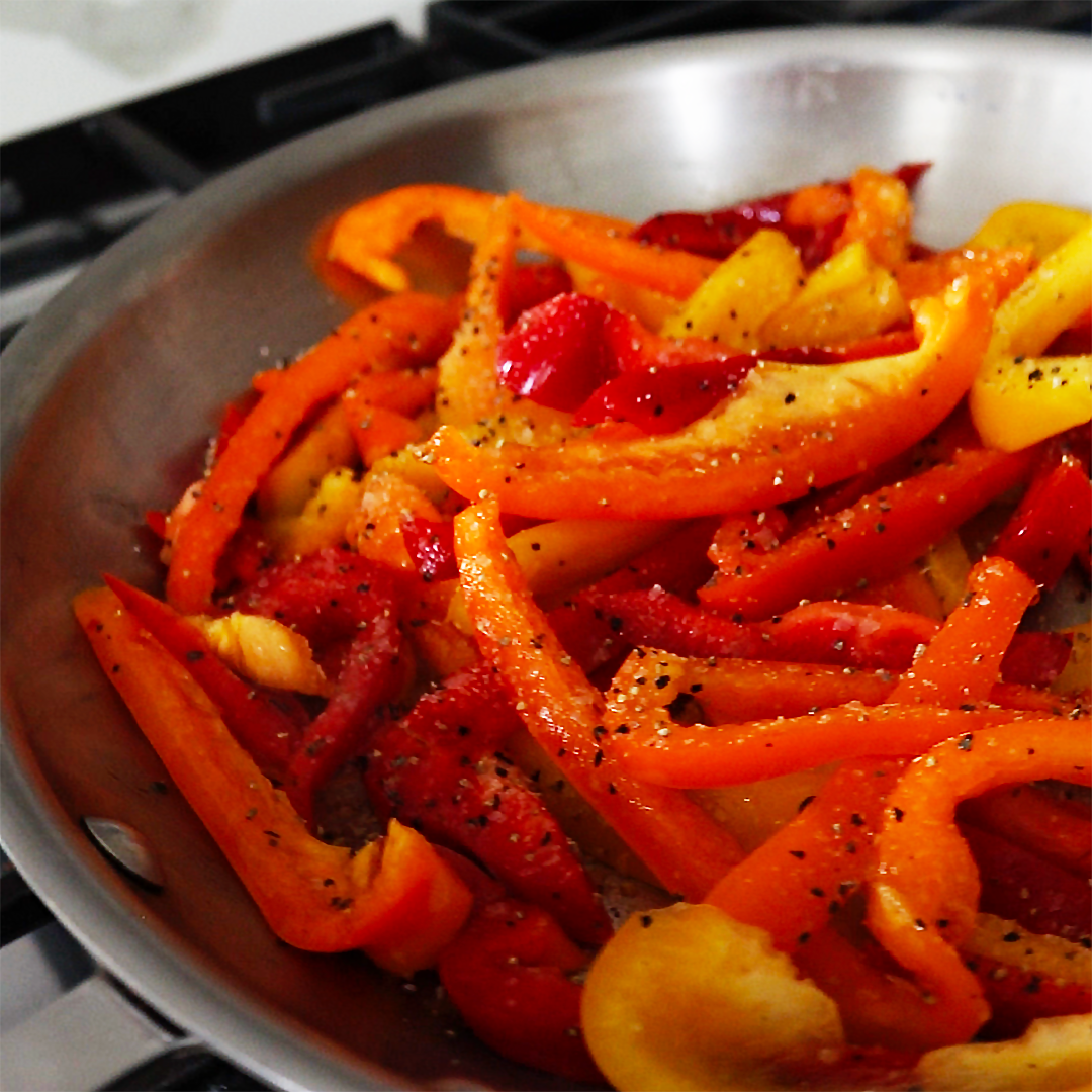 Sauteed red, orange, and yellow bell pepper strips in a stainless skillet, sprinkled with cracked black pepper
