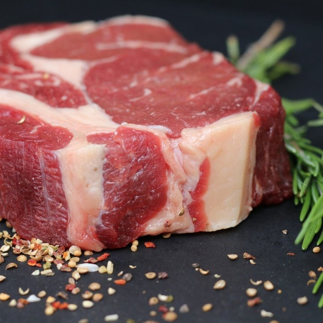 Raw ribeye steak with visible marbling on a black surface, chili flakes scattered and a rosemary sprig.