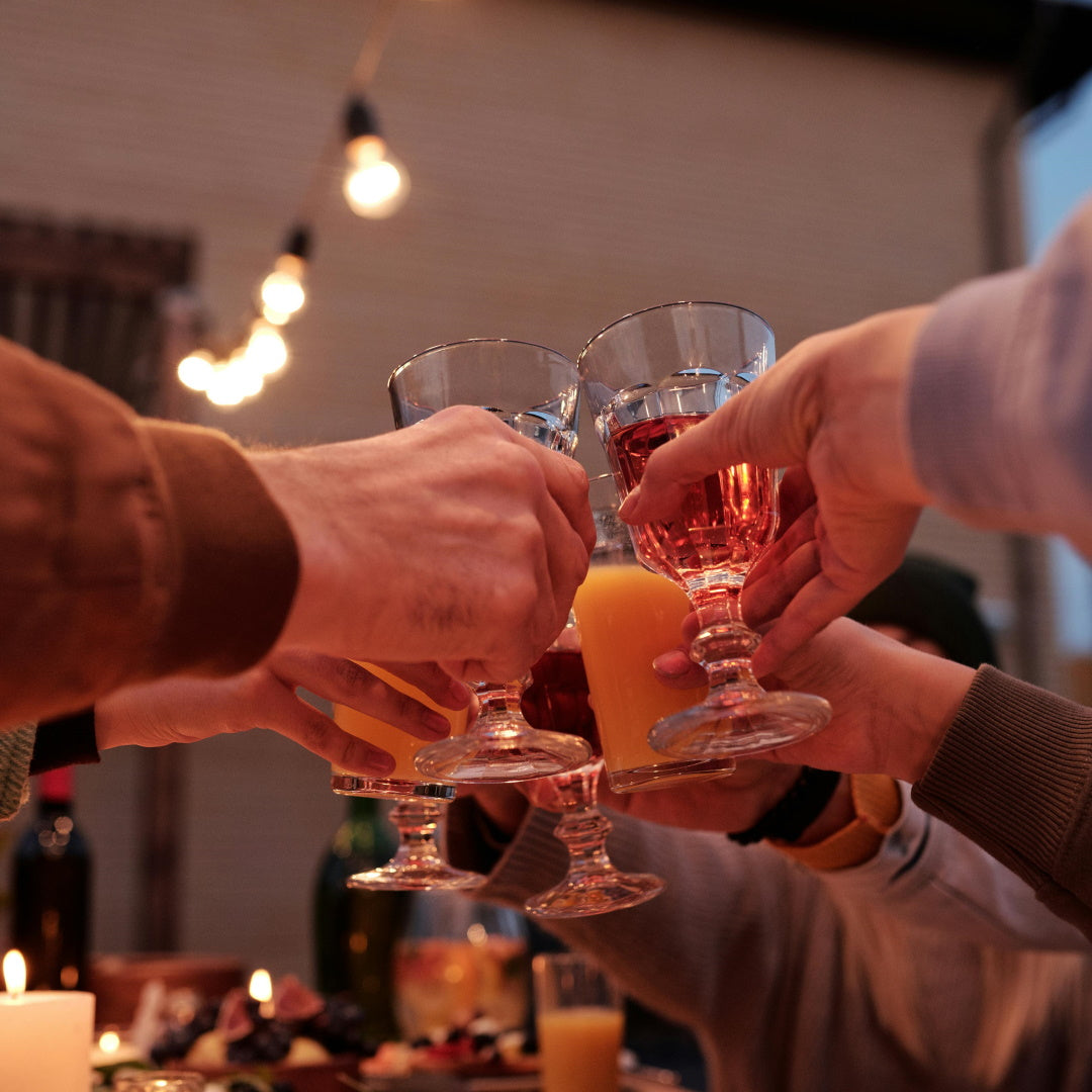 Friends clinking stemmed glasses of wine and orange juice over a candlelit holiday dinner table.