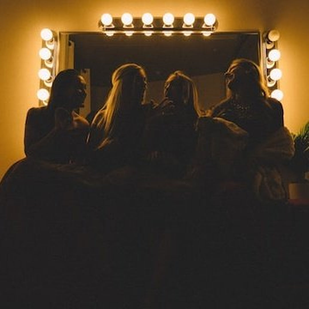 Four women silhouetted on a couch before a lighted dressing mirror with round bulbs, warm amber backlight