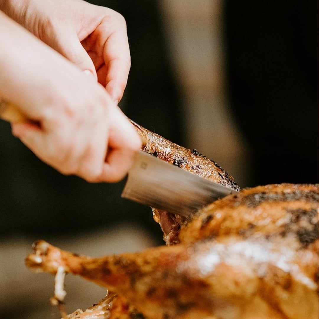 Hands carving a herb-buttered roast turkey, browned skin visible as a cleaver slices the leg.