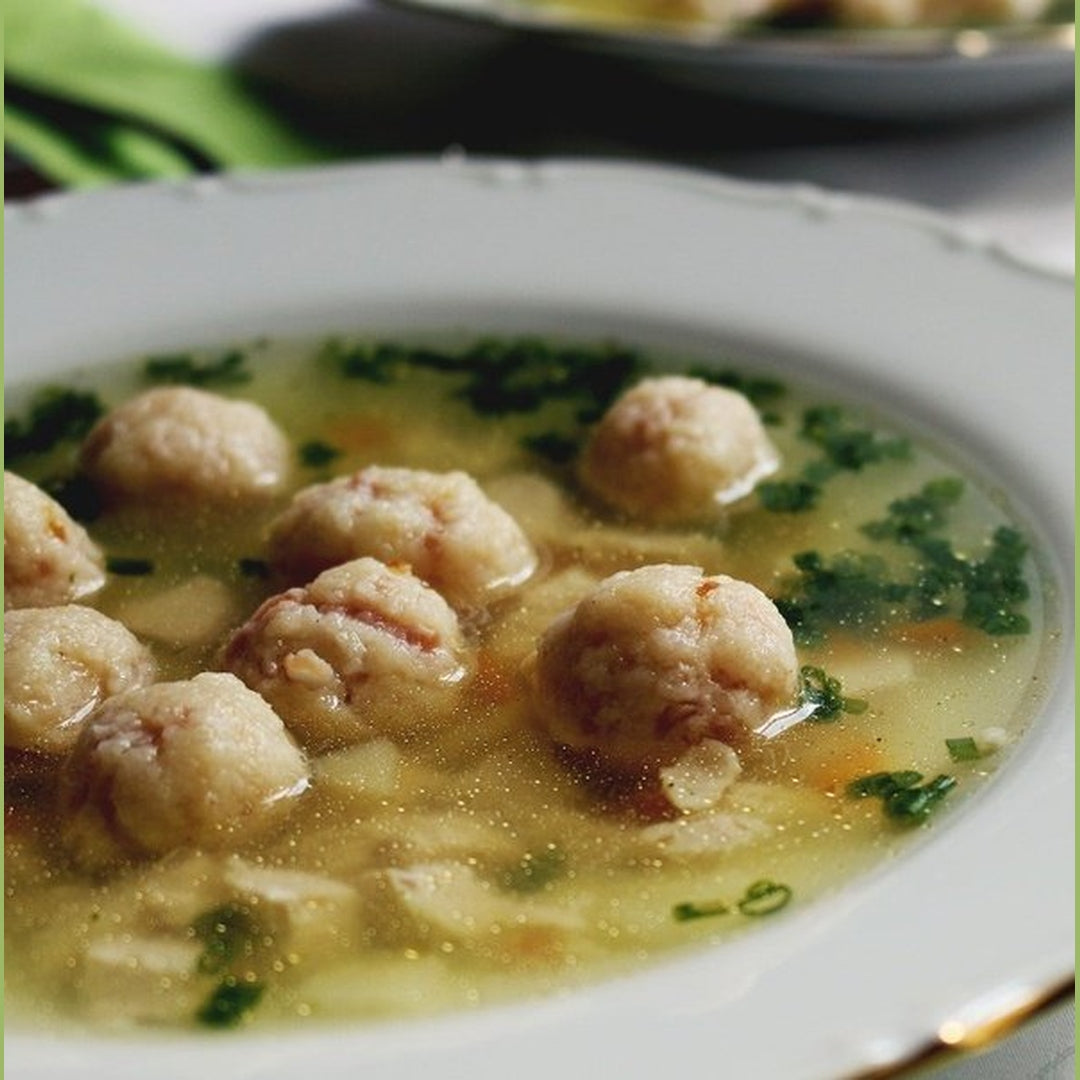 Bowl of clear chicken broth with several matzoh balls and chopped parsley in a white rimmed soup plate.