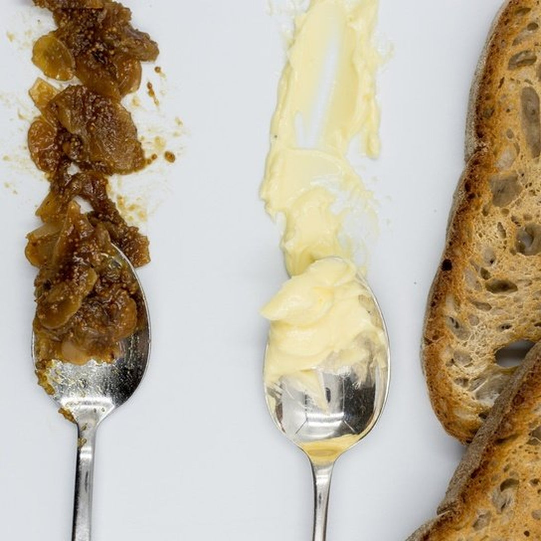 Two spoons with a caramelized onion spread and softened butter beside toasted bread slices on a white surface
