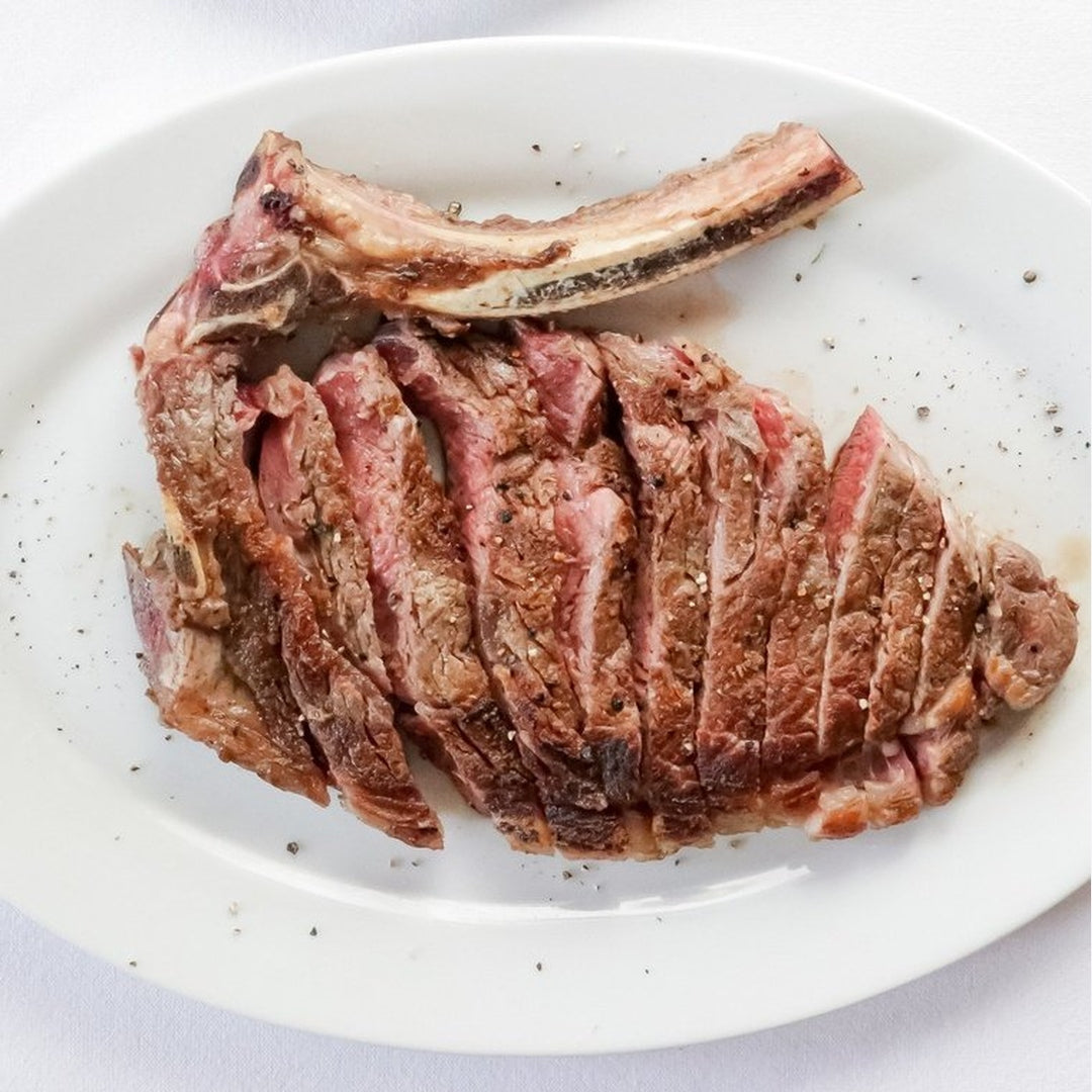 Sliced medium-rare ribeye with bone, lightly seasoned with Sergio's Seasoning on a white oval plate.