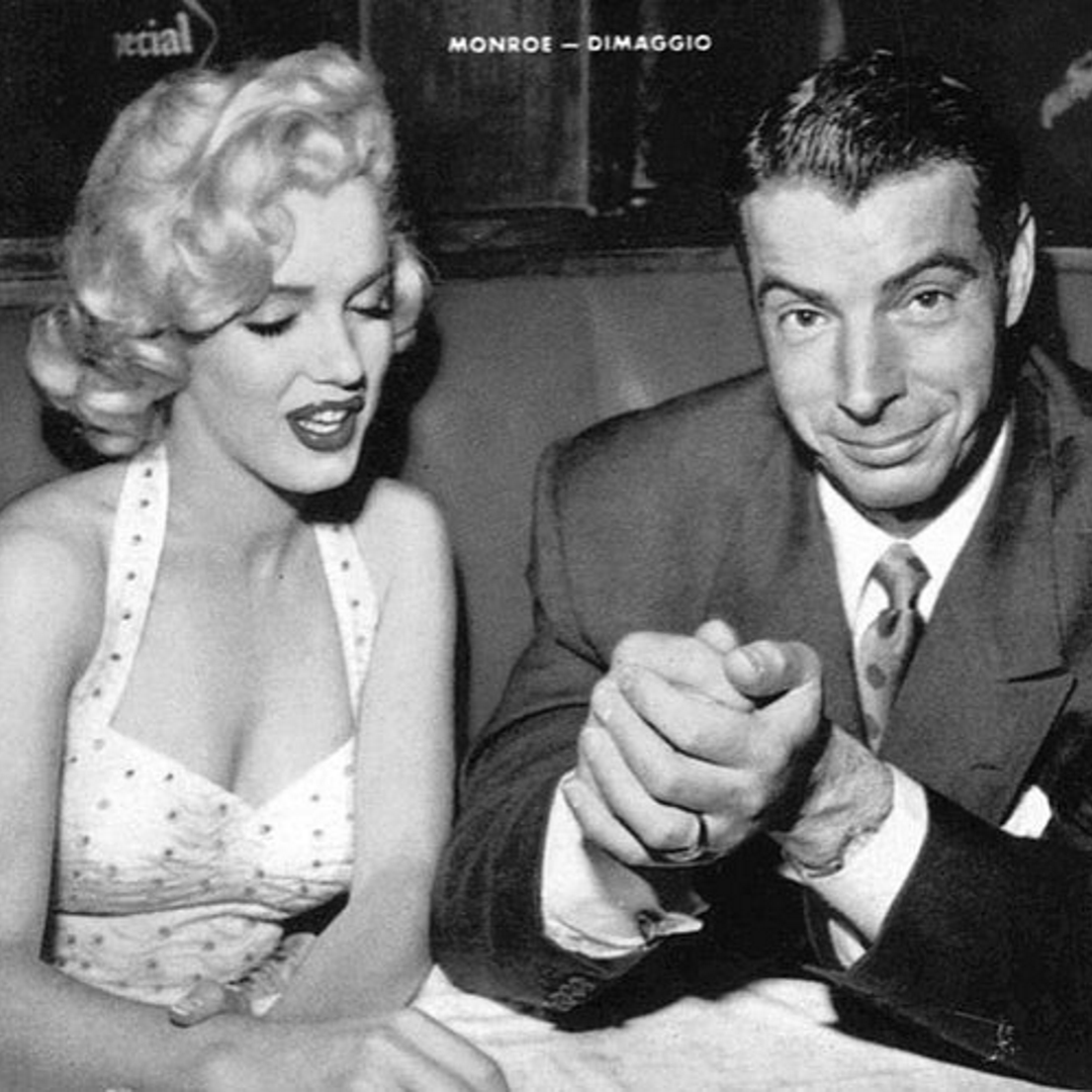 Marilyn Monroe in a halter dress smiling beside Joe DiMaggio in a suit with clasped hands, seated together in a restaurant booth.