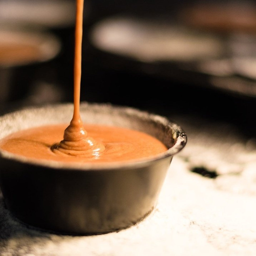 Metal ramekin being filled with smooth golden-orange aioli as a thin stream pours into the center.
