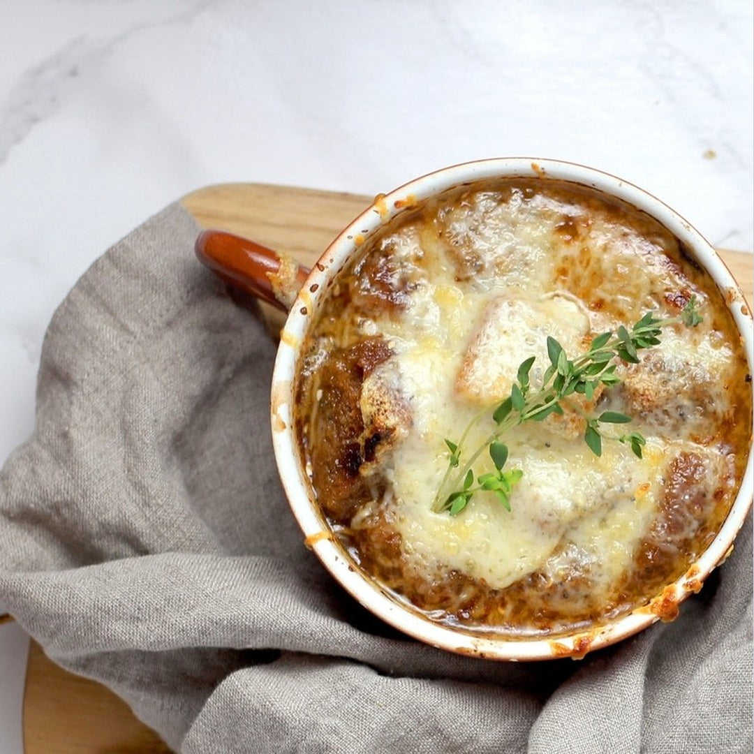 Bowl of steakhouse French onion soup topped with melted cheese, toasted bread and a thyme sprig on a linen napkin.