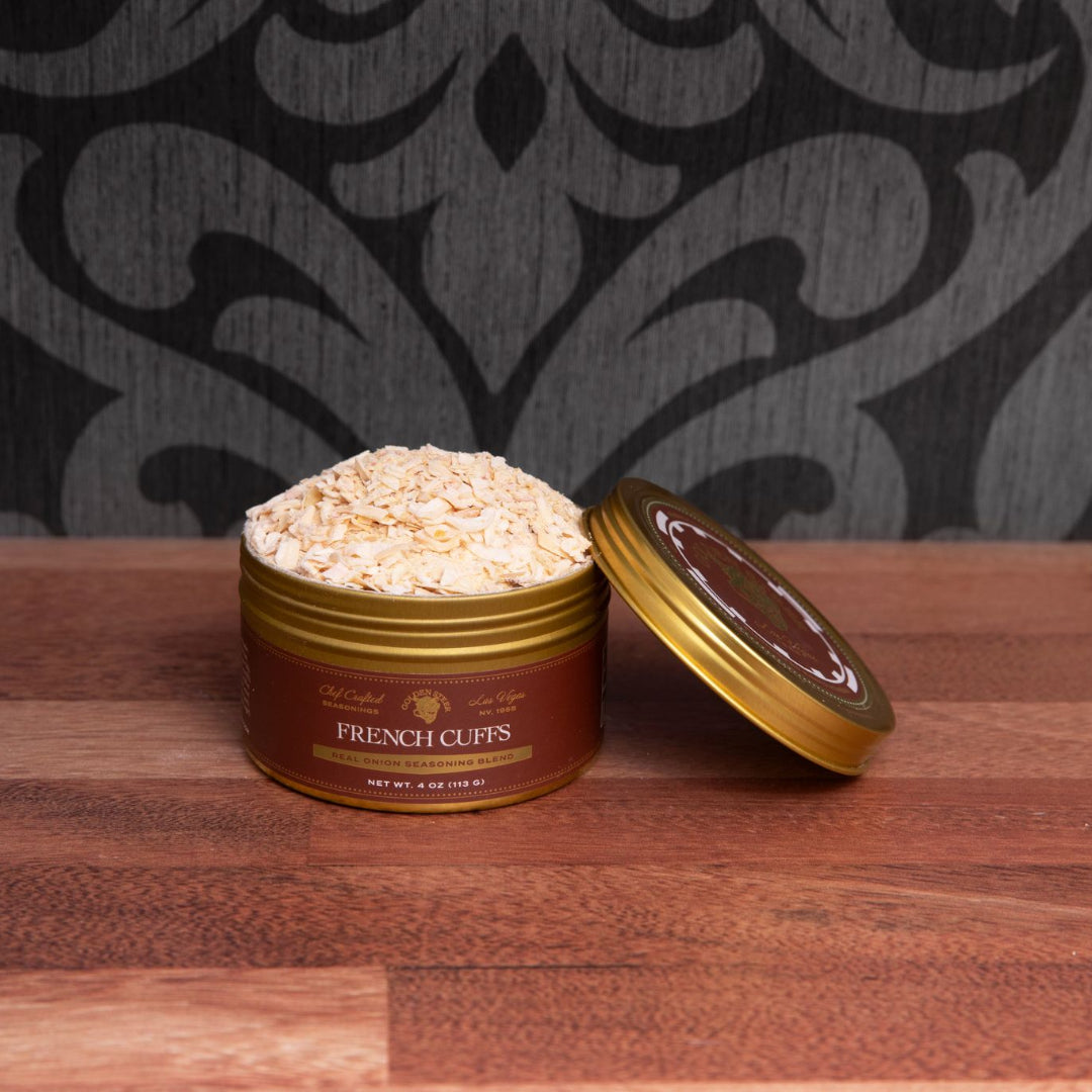Front view of French Cuffs dried onion seasoning tin opened to show pale dried onion flakes on a wood countertop against a dark patterned wall