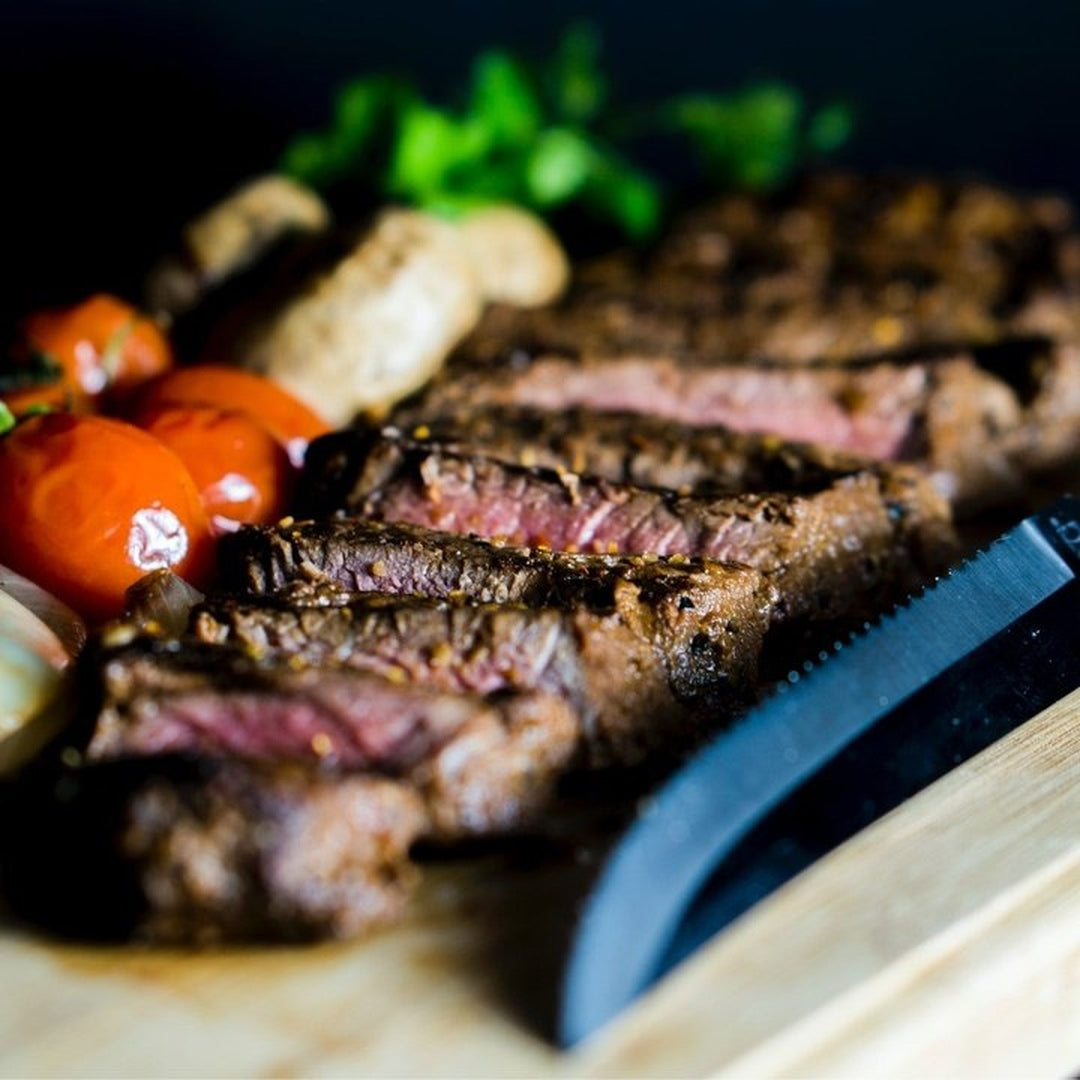 Sliced medium-rare steak on a wooden board with roasted cherry tomatoes, mushrooms and a serrated knife