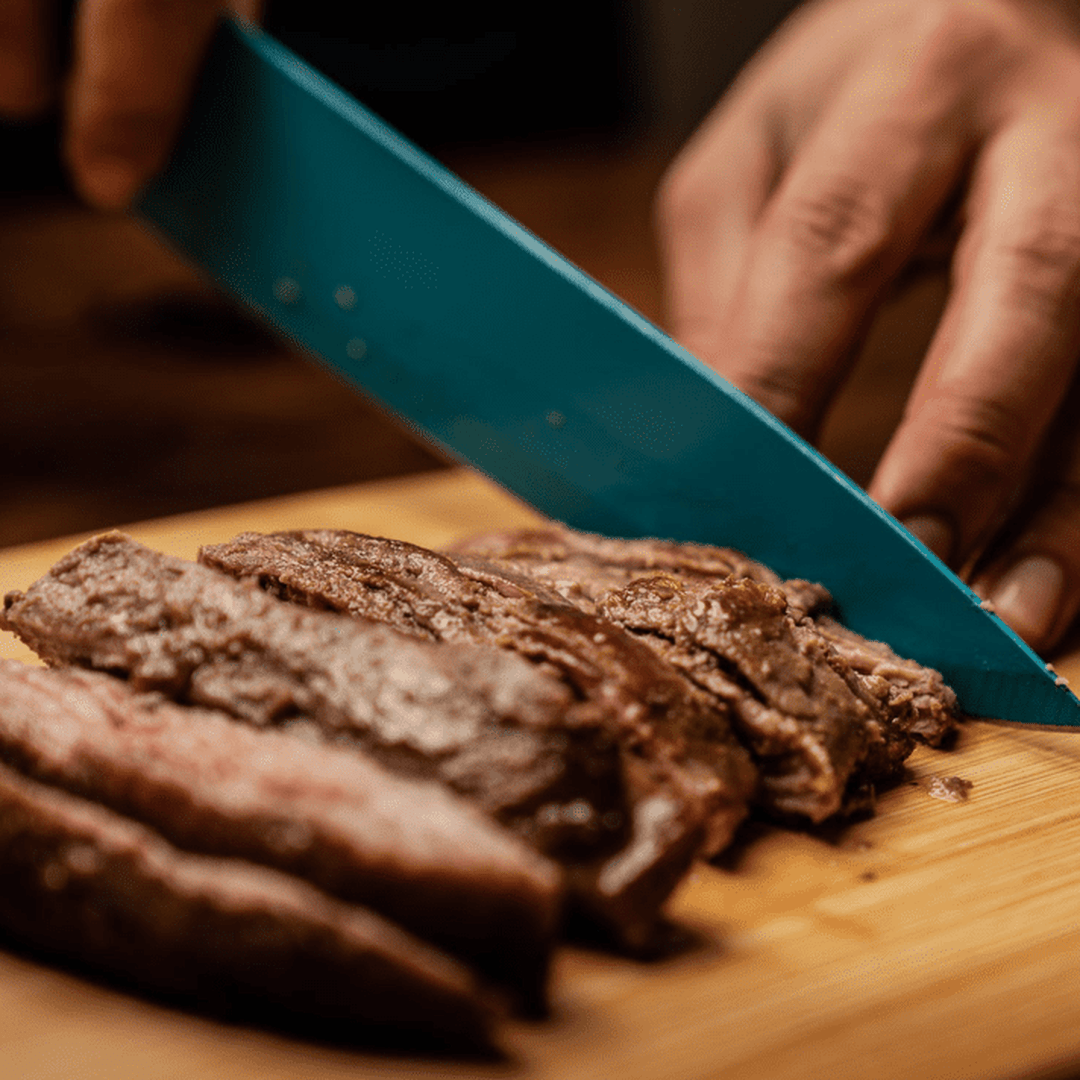 Hand slicing cooked steak with a teal chef's knife on a wooden cutting board
