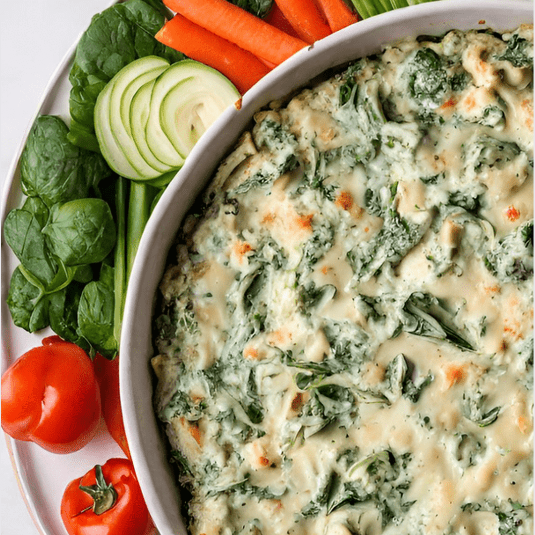 Baked spinach and artichoke dip with melted cheese in a white baking dish, plated with spinach leaves, cherry tomatoes and carrot sticks.