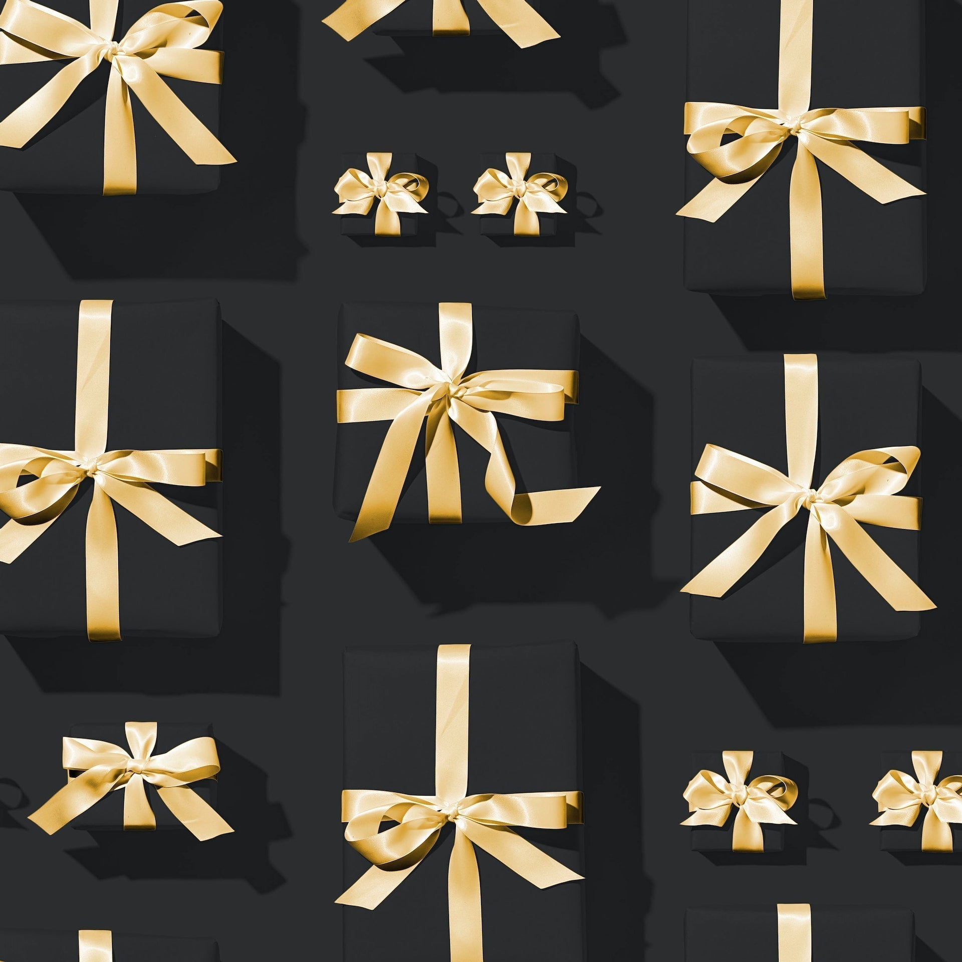 Black gift boxes with gold satin ribbons arranged in a grid on a dark background