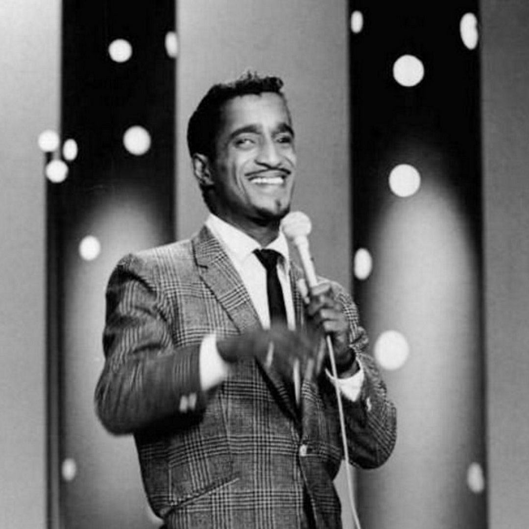 Sammy Davis Jr. in a checkered suit holding a microphone on stage with round light spots in the background