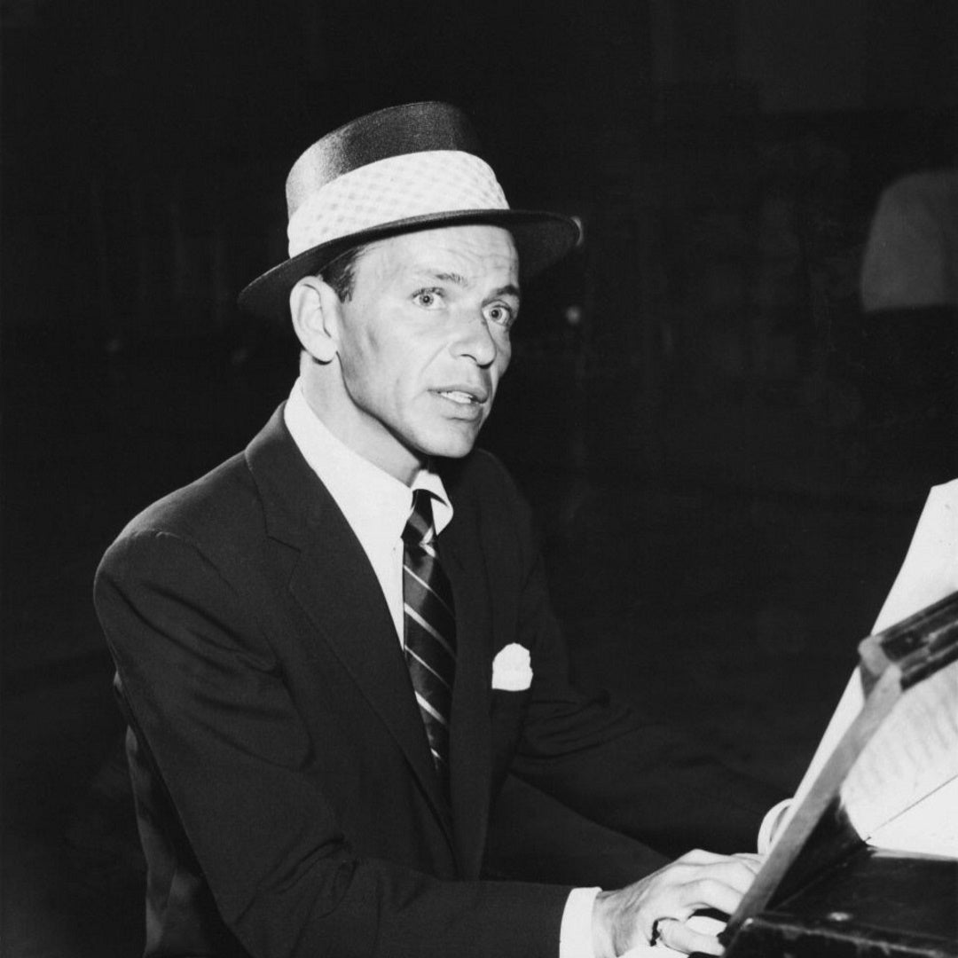 Frank Sinatra in a suit and boater hat playing piano and looking toward the camera in a dimly lit room