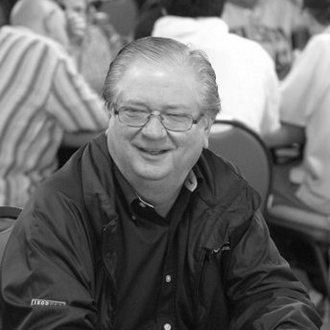 Smiling older man wearing glasses and a dark jacket, seated at a crowded table in a busy event room.
