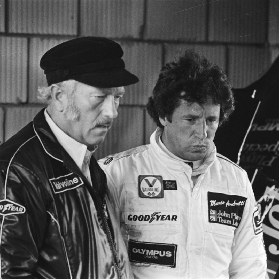 Colin Chapmann and Mario Andretti pictured in racing jackets, patches read Valvoline; GOODYEAR; OLYMPUS CAMERAS; Mario Andretti; John Player Team