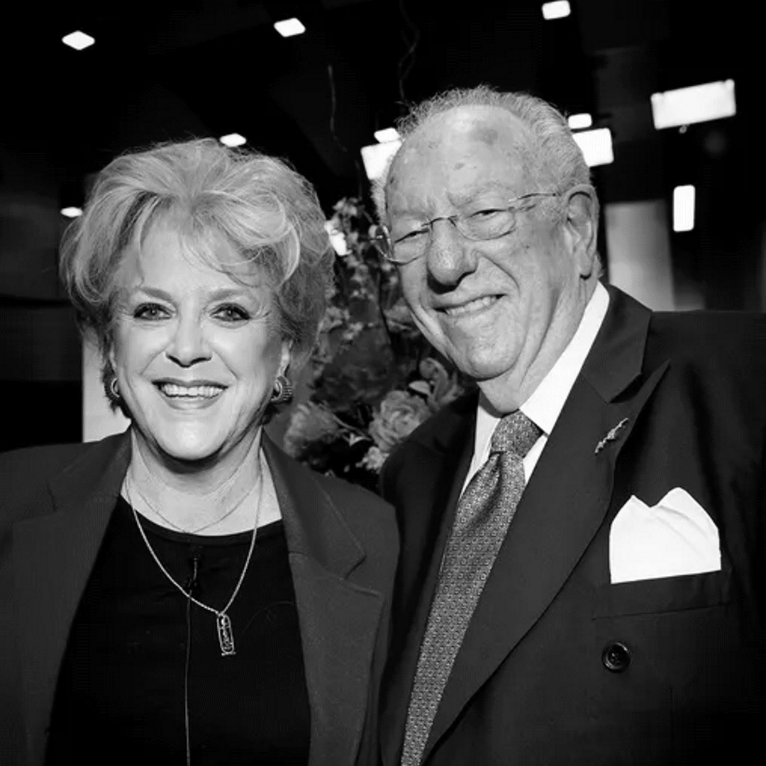 Carolyn and Oscar Goodman pictured smiling at an indoor event, man in a suit and tie with pocket square and woman wearing a necklace, black-and-white photo