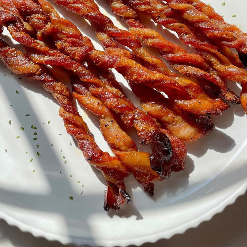 Crispy twisted bacon strips arranged on a white plate with sprinkled herbs and sunlight