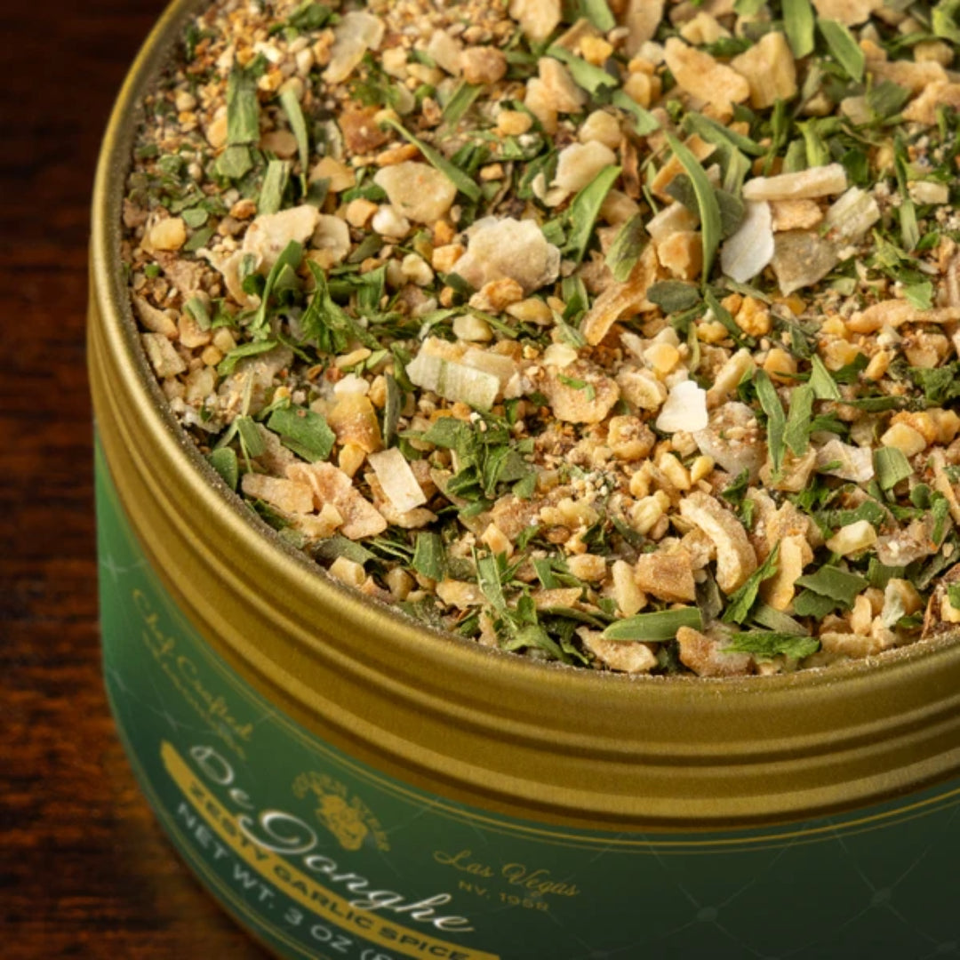 Close-up of De Jonghe seasoning tin showing garlic flakes, chopped herbs, and spices in an open gold tin on wood