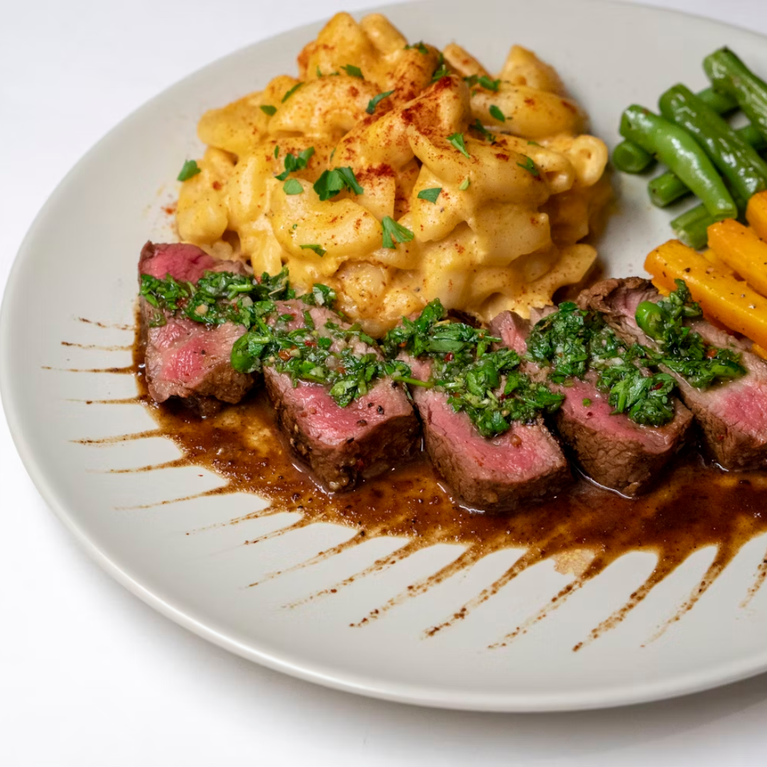 Sliced medium-rare steak with chimichurri, macaroni-and-cheese, green beans and carrots on a white plate.