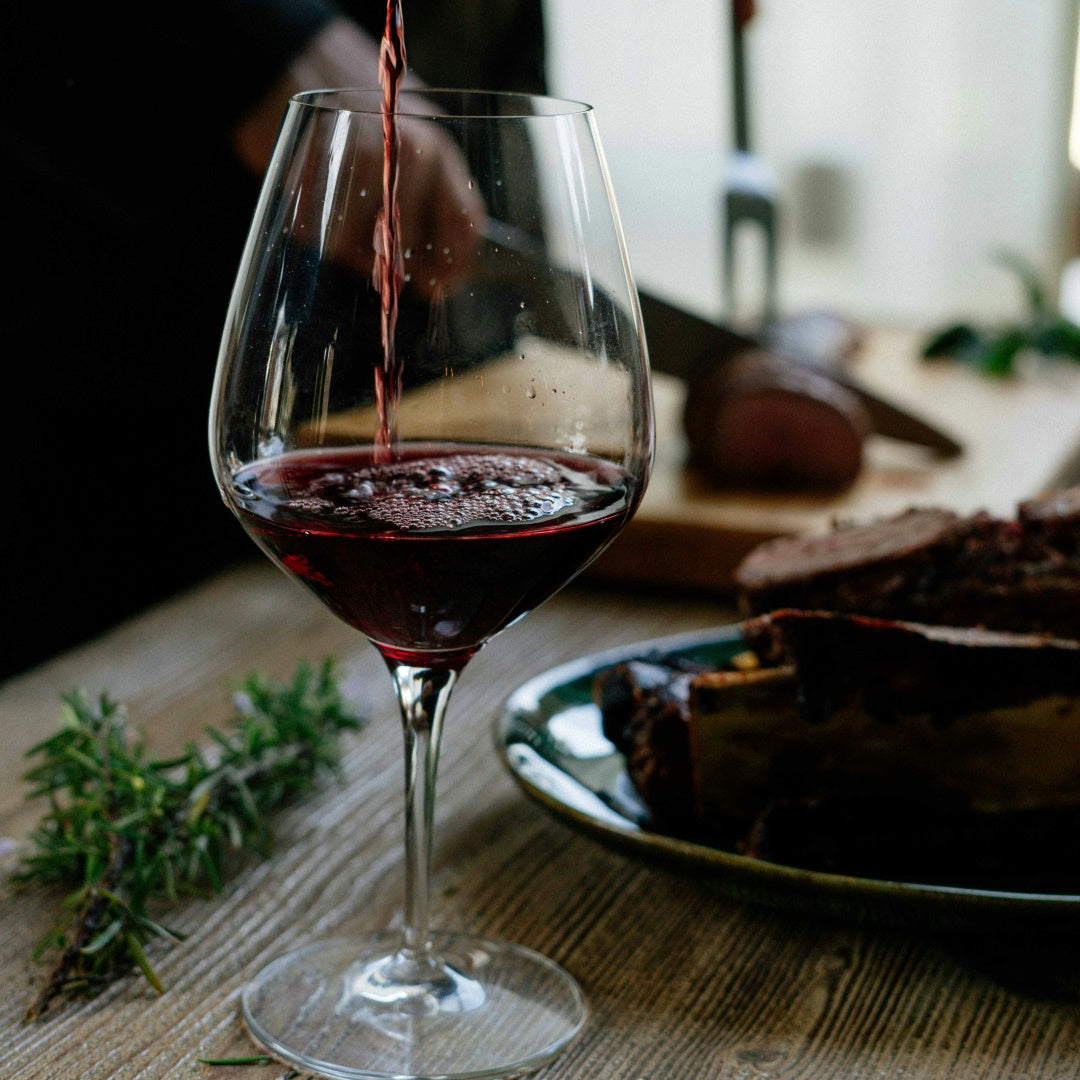 A glass of red wine being poured beside a plate of steak ribs and sprigs of rosemary on a wooden table