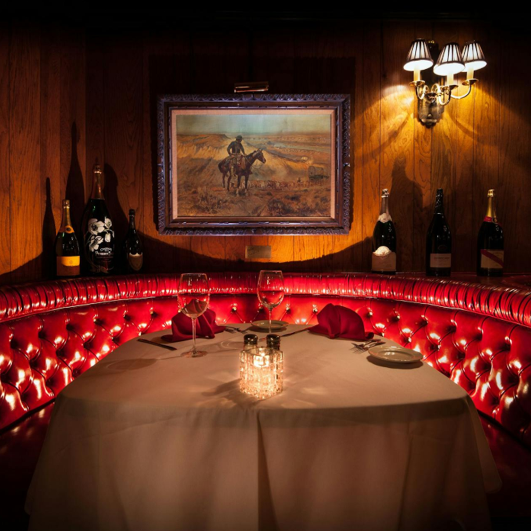 Red tufted booth for two with white tablecloth, candle, wine glasses, cowboy painting and champagne bottles