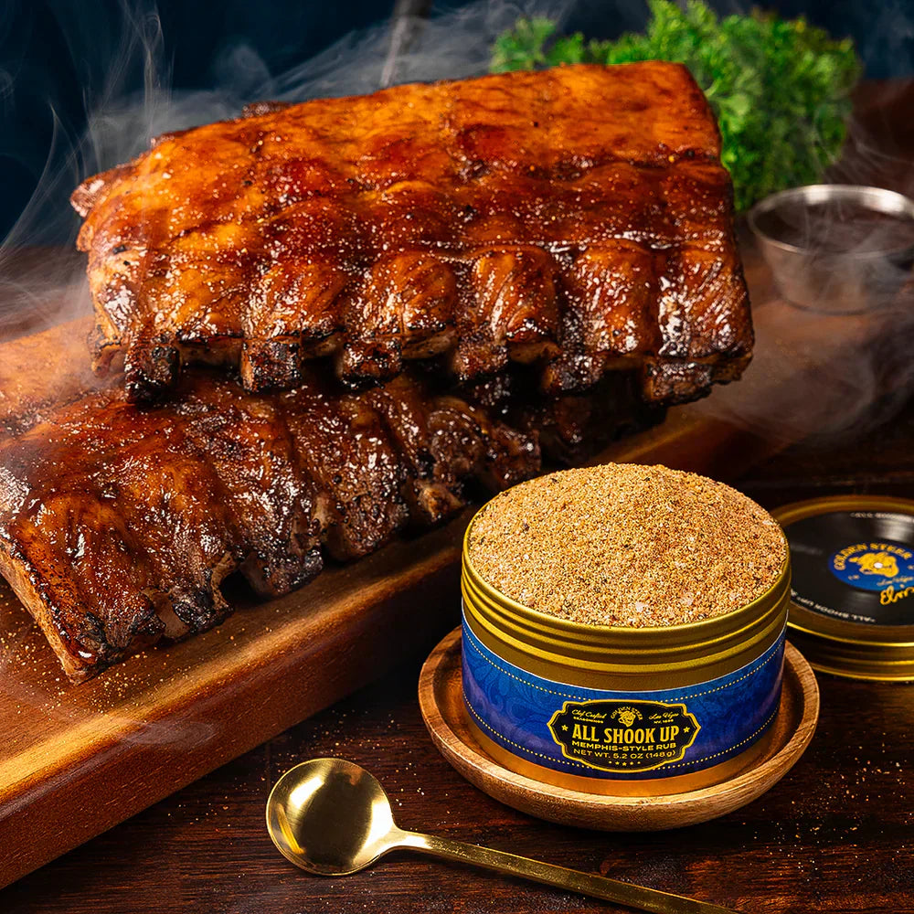 Smoked pork ribs on a board with a tin of seasoning labeled 'ALL SHOOK UP' 'MEMPHIS-STYLE RUB' and a gold spoon.