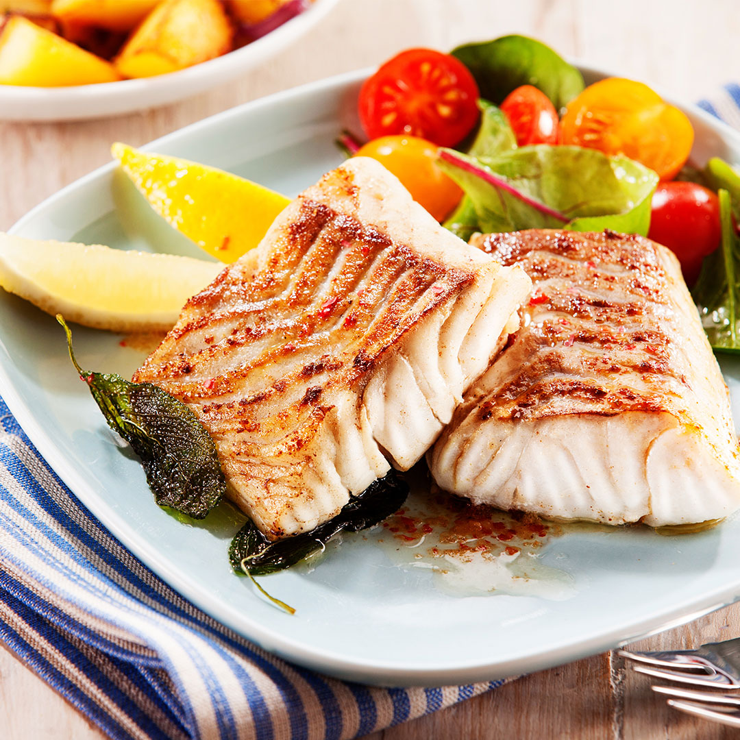 Grilled fish fillets with char marks on a plate, lemon wedges, mixed cherry tomato salad and fried sage