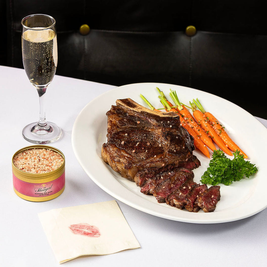 Seared, sliced ribeye with roasted baby carrots on a white plate, with a Showgirl seasoning tin, champagne flute, and napkin with a lipstick kiss