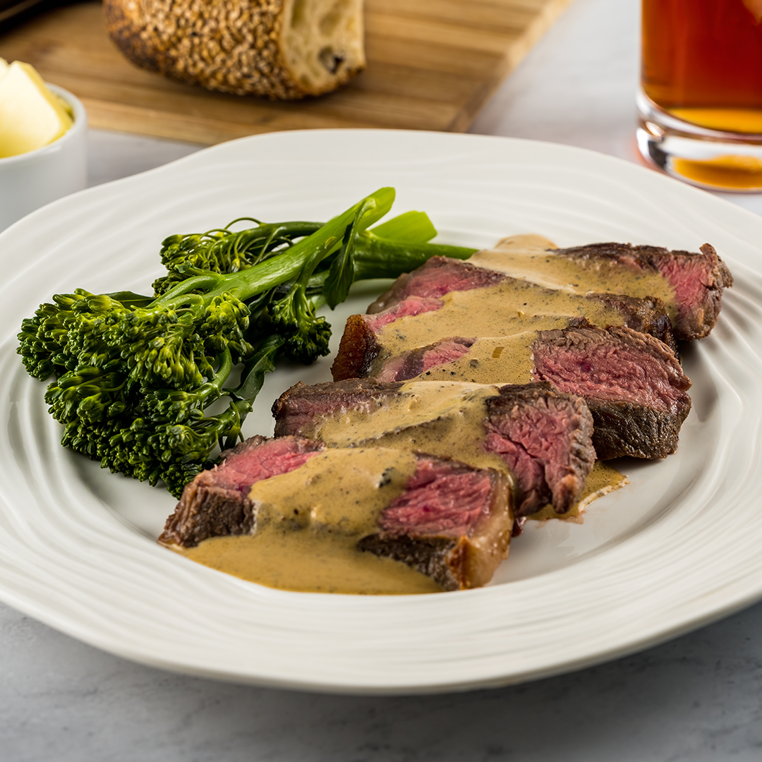 Slices of peppercorn steak au poivre with creamy sauce and a side of broccolini on a white plate