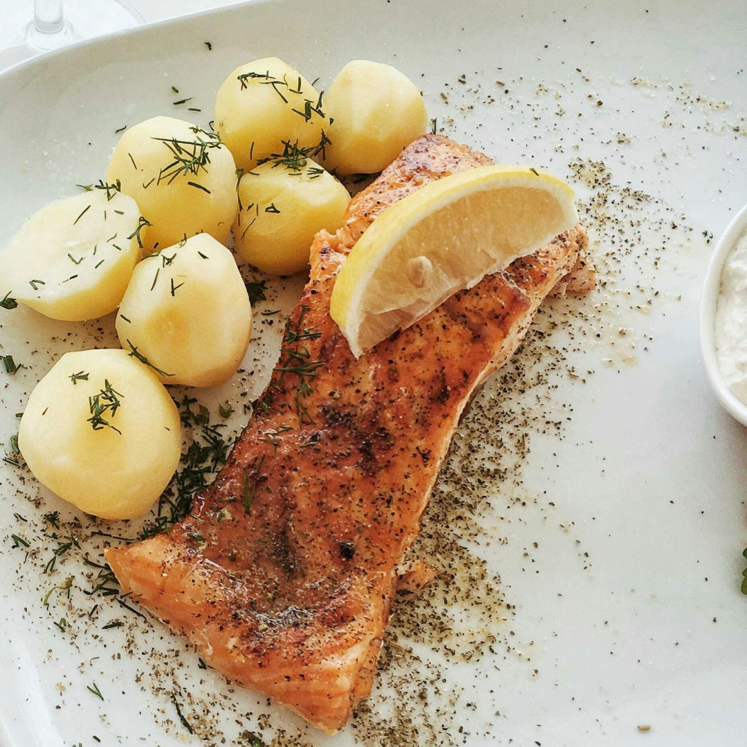 Baked salmon fillet with a lemon wedge served alongside boiled potatoes and dill on a white plate.