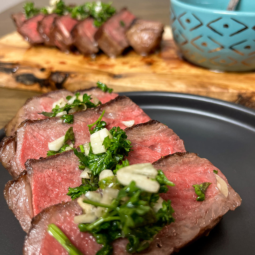 Sliced New York strip steak with herb chimichurri on a dark plate