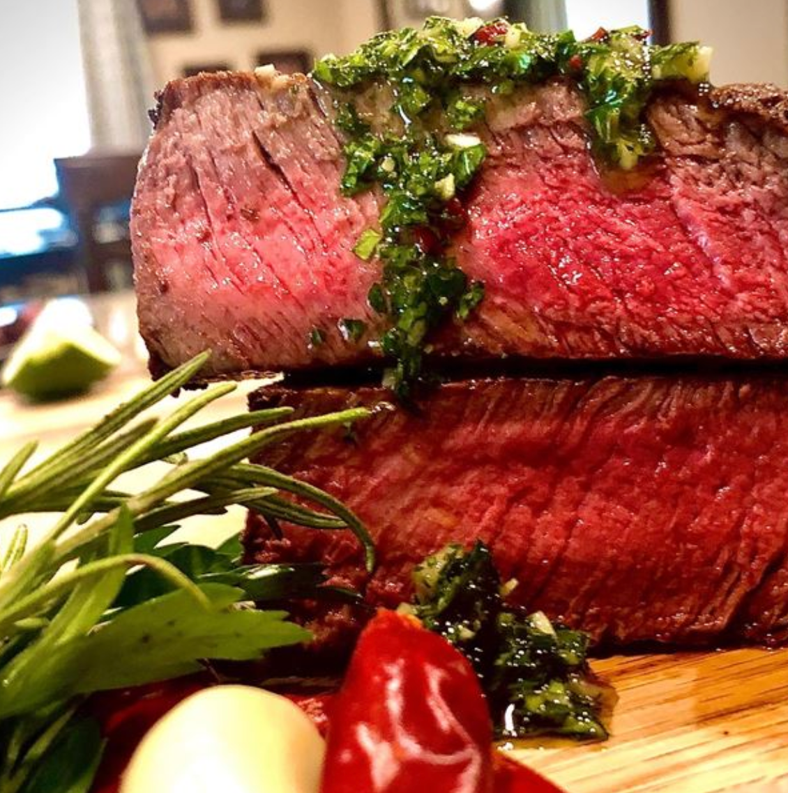 Thick filet mignon topped with chimichurri on a wooden board with herbs in the foreground.