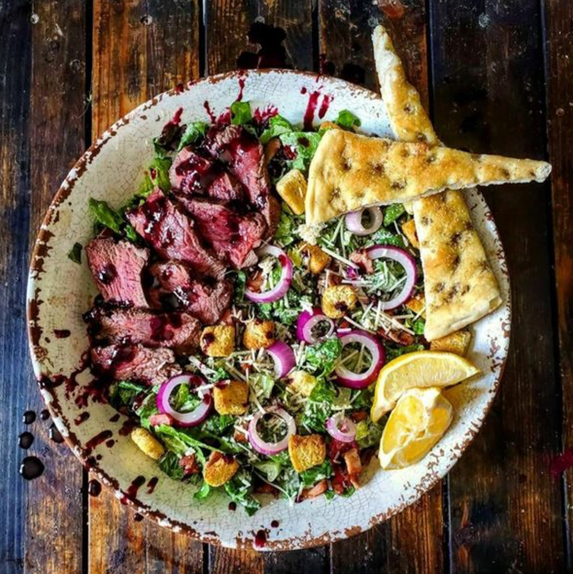 USDA Prime ribeye on a Caesar salad with greens, red onions, croutons, lemon wedges, and triangular flatbread.
